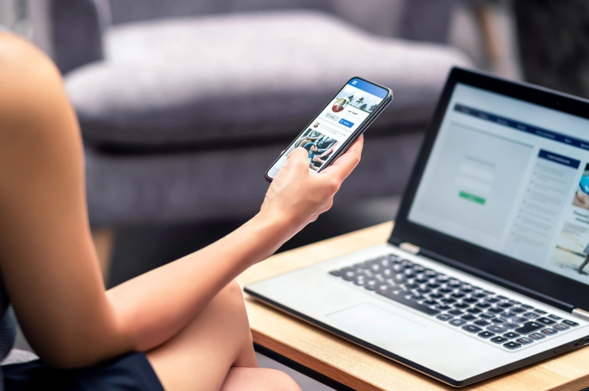 Person sitting with a laptop on a table and a smartphone in hand, browsing Facebook.