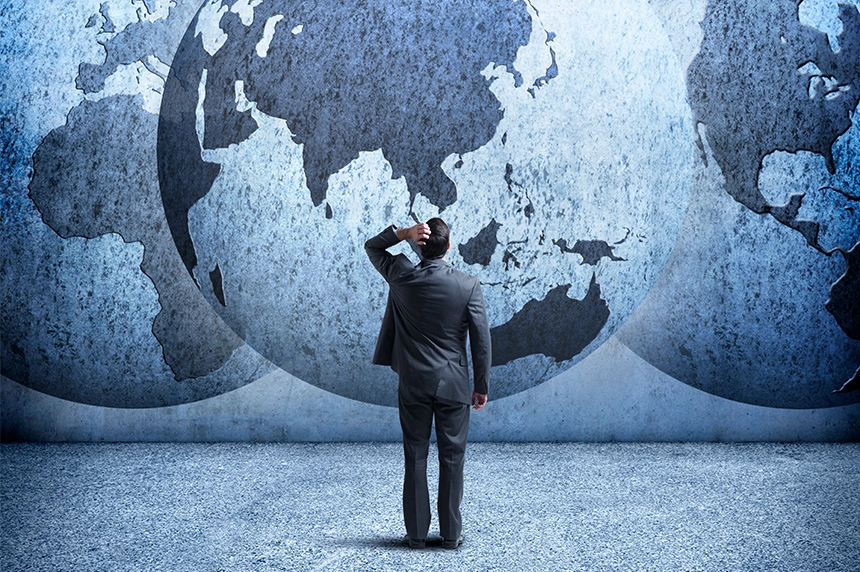 A person in a suit standing in front of a large world map depicted on a textured wall, looking thoughtfully at the map.