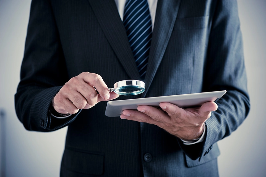 A person in a business suit uses a magnifying glass to examine a tablet.